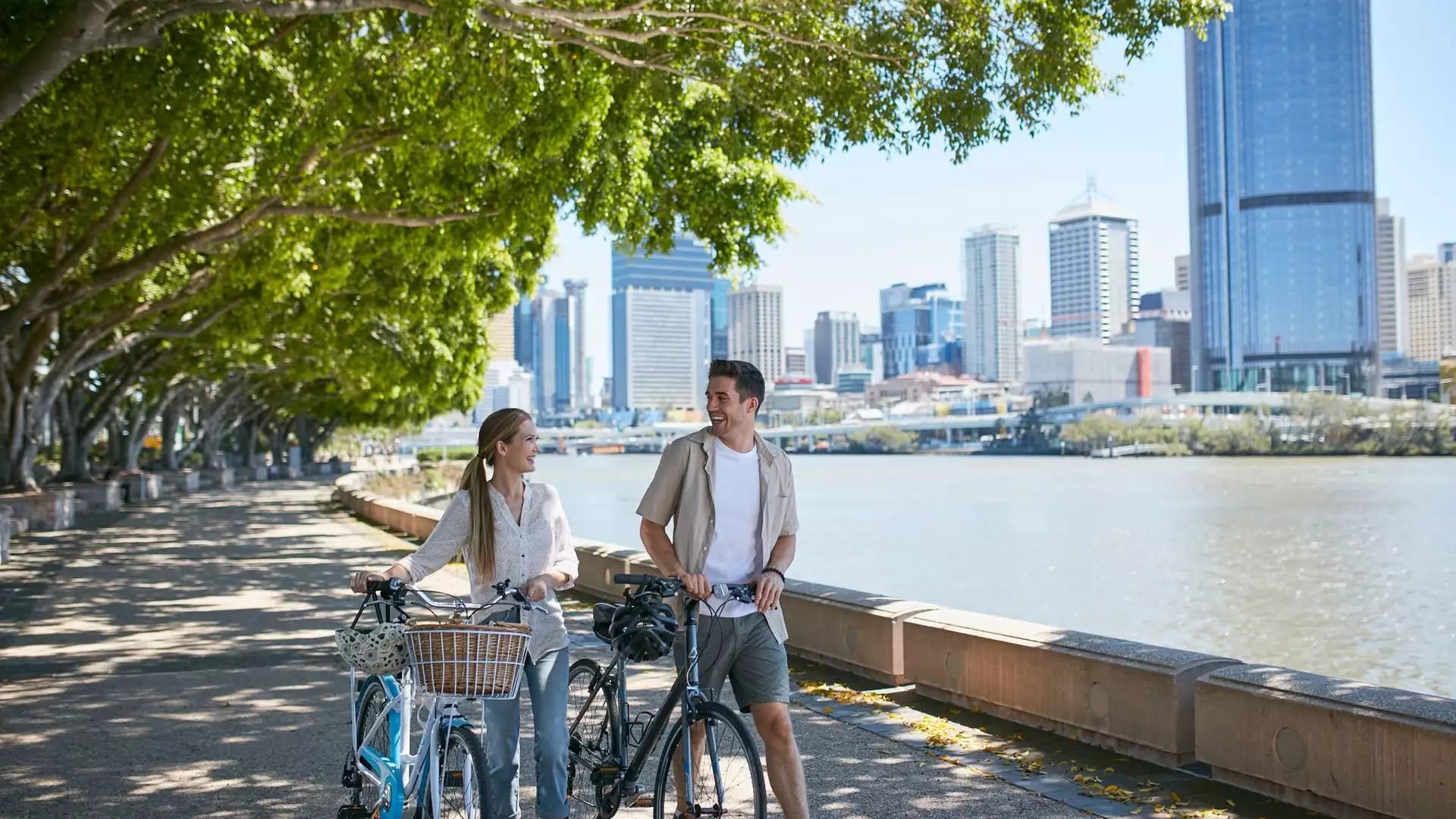 Man and woman walking with bicycles along a tree-lined riverside path with Brisbane city buildings in the background.