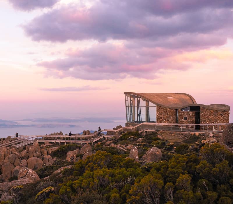 A modern stone and glass lookout building on a rocky mountain summit overlooking a coastal city under a pink sunset sky in Tasmania.