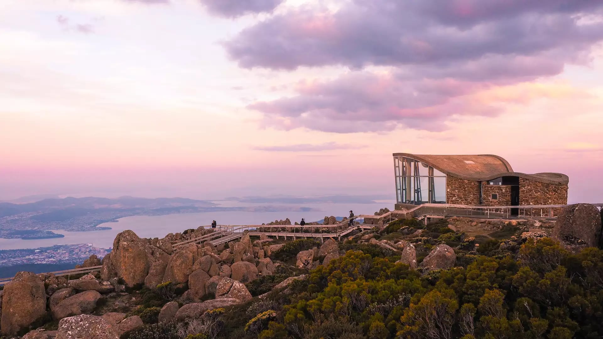 A modern stone and glass lookout building on a rocky mountain summit overlooking a coastal city under a pink sunset sky in Tasmania.