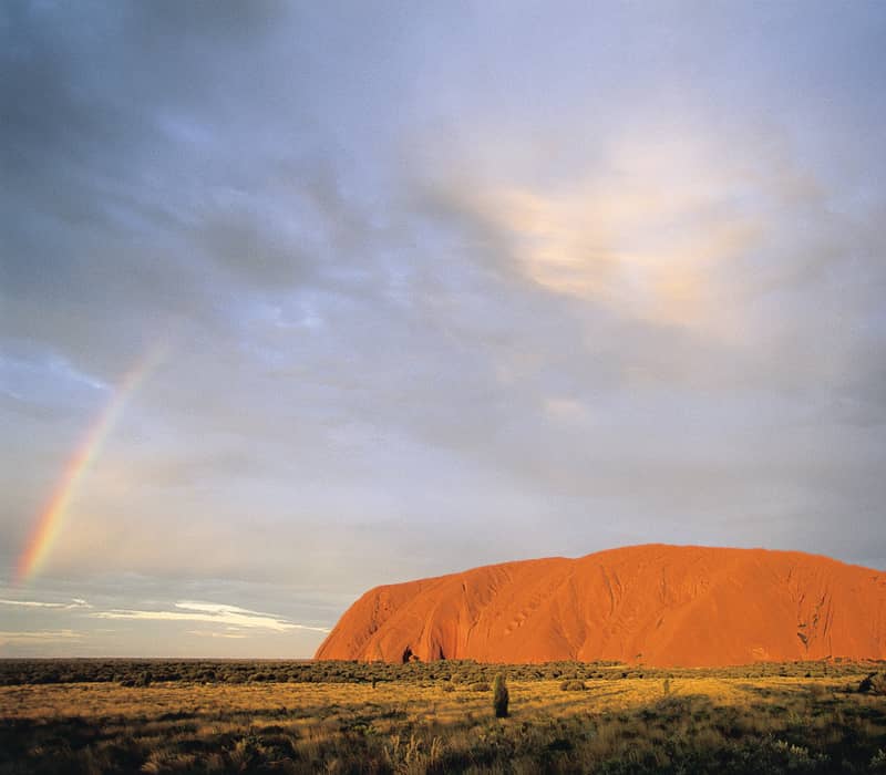 The red monolith of Uluru in the distance with a bright rainbow appearing on the left side under a cloudy sky.