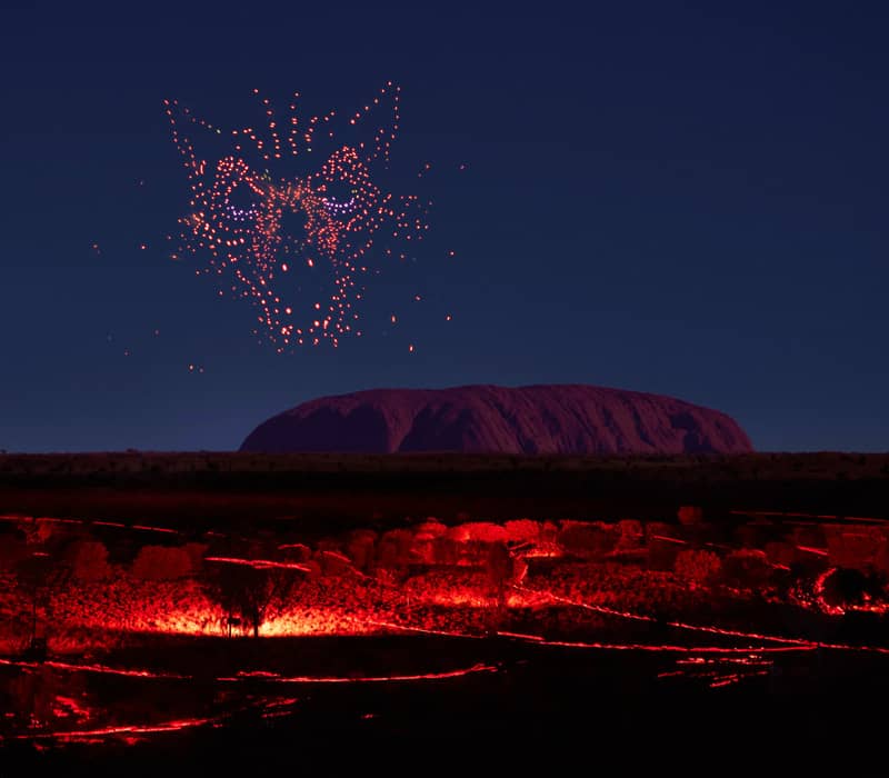 Vertical night shot of a red glowing drone formation shaped like a face in the sky above the dark silhouette of Uluru and lit desert paths.
