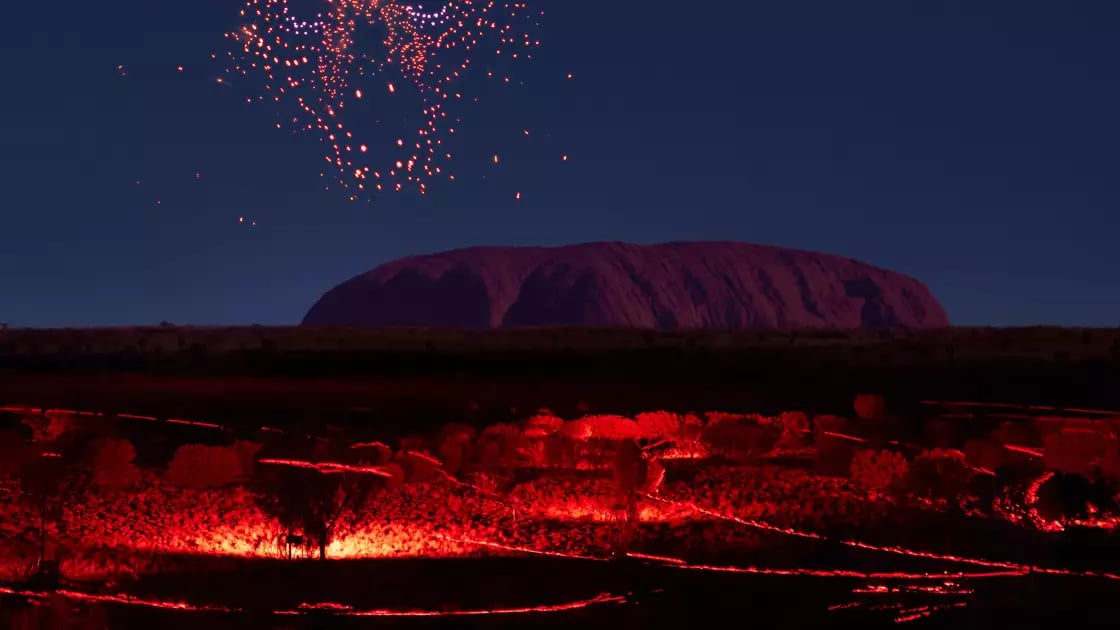 Vertical night shot of a red glowing drone formation shaped like a face in the sky above the dark silhouette of Uluru and lit desert paths.