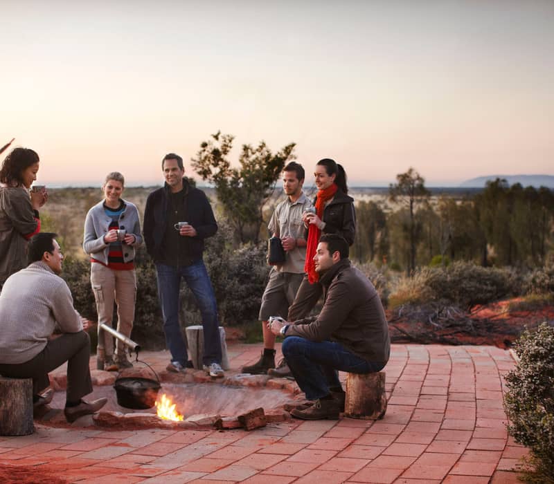 A small group of people standing around a desert campfire at dawn with Uluru visible on the horizon.