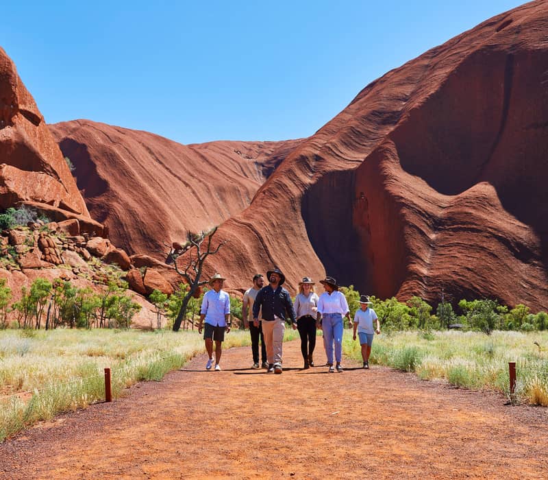 A guided group of hikers walking along the red earth path at the base of the Uluru monolith under a clear blue sky.
