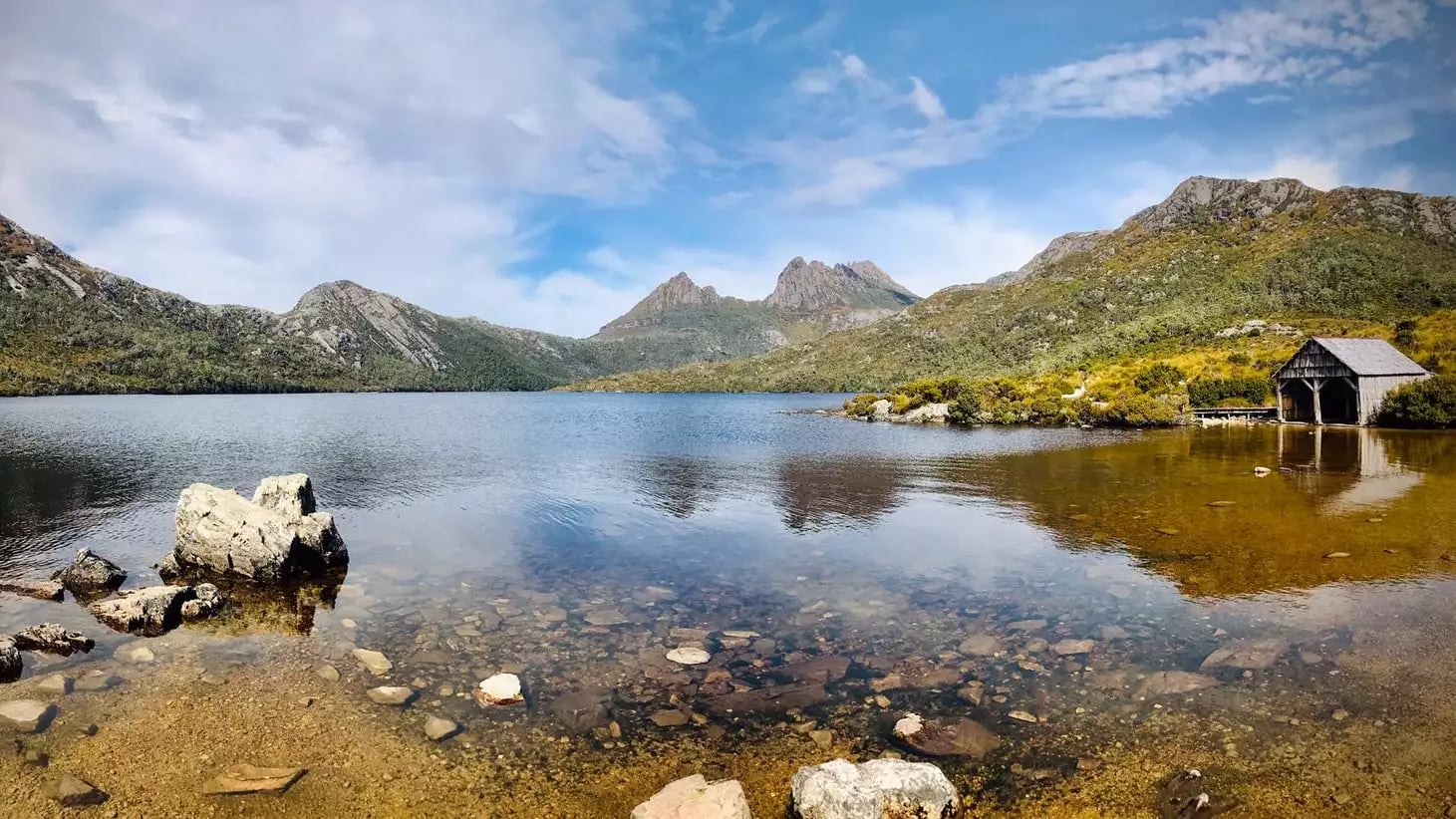 A wide landscape view of Dove Lake in Tasmania featuring the historic wooden boatshed and Cradle Mountain under a blue sky.