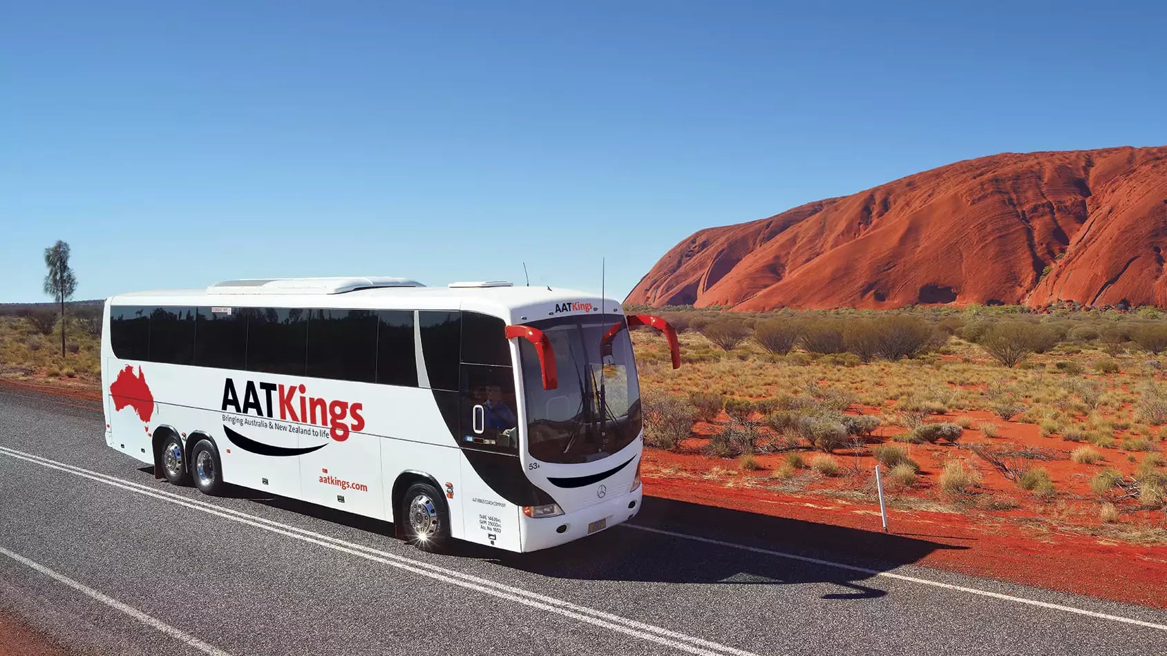 A white AAT Kings tour bus driving on a paved road through the red desert with Uluru looming on the horizon.