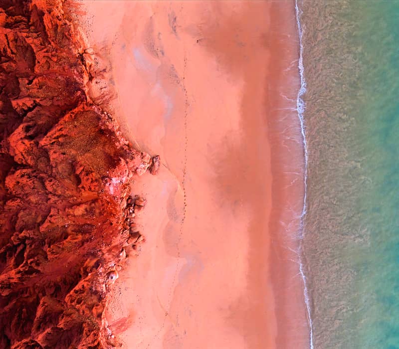 A high-angle aerial shot showing a deep red, jagged cliff line contrasting sharply against a bright turquoise ocean with small white waves.
