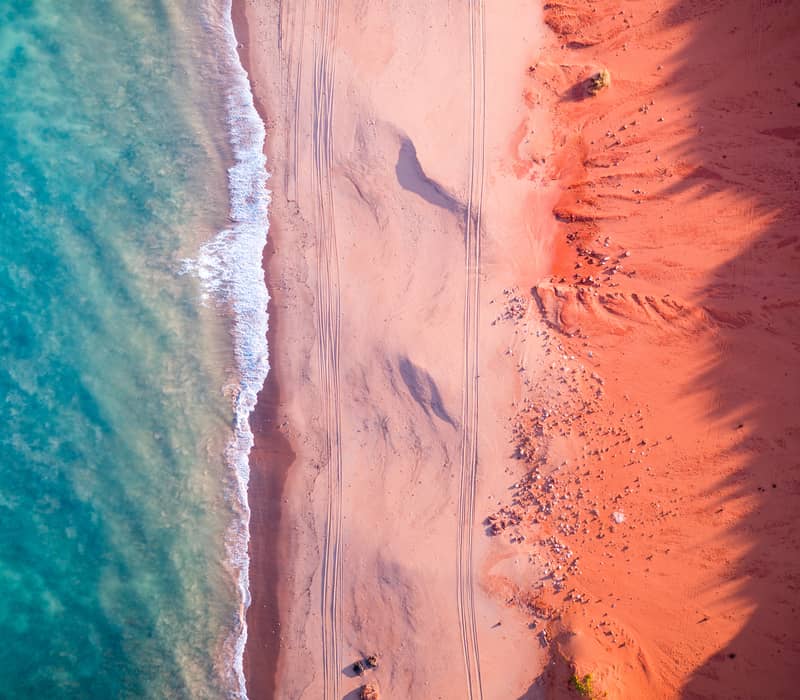 A high-angle aerial shot showing a deep red, jagged cliff line contrasting sharply against a bright turquoise ocean with small white waves.