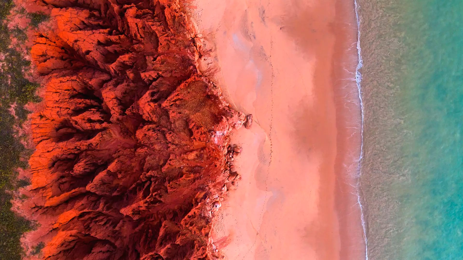 A high-angle aerial shot showing a deep red, jagged cliff line contrasting sharply against a bright turquoise ocean with small white waves.