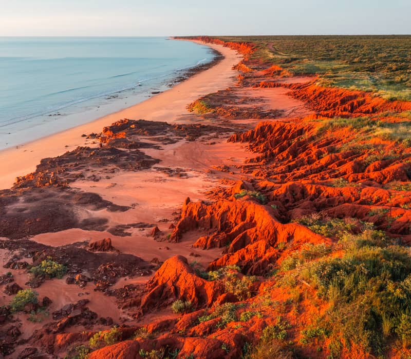 A high-angle aerial shot showing a deep red, jagged cliff line contrasting sharply against a bright turquoise ocean with small white waves.