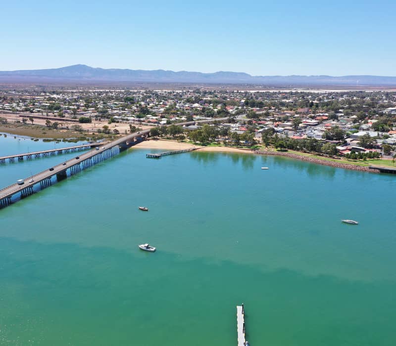 Aerial perspective of a long coastal bridge crossing green-blue water leading into a town with mountain ranges on the horizon.