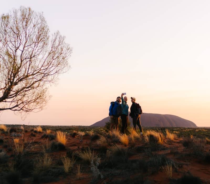 Three people stand on a sandy ridge at dawn, smiling for a phone photo with the massive, dark rounded shape of Uluru visible on the horizon behind them.