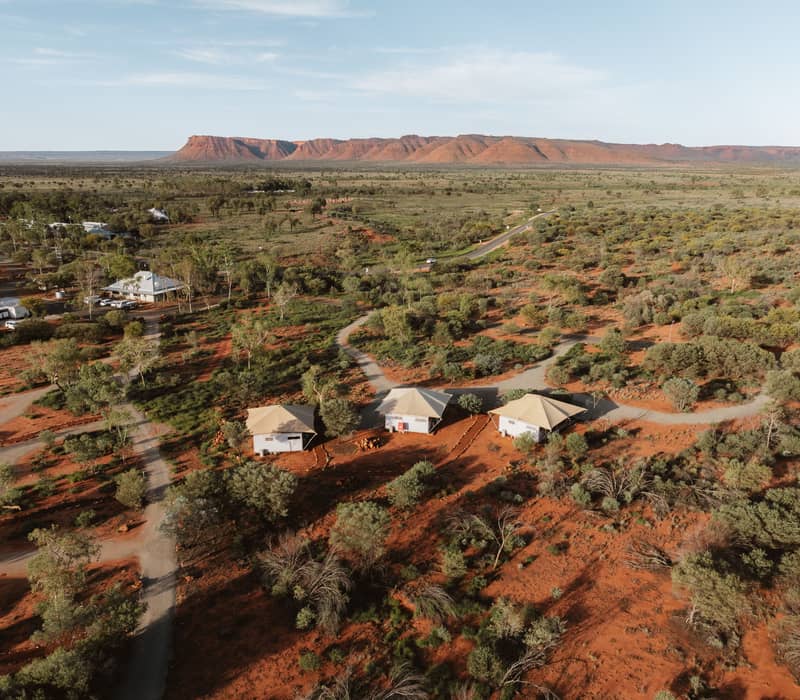 A high-angle drone view shows three modern safari-style glamping tents with tan roofs arranged on bright red earth among green desert trees.