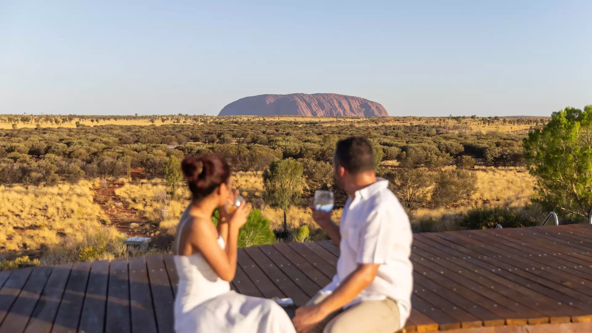 A man and woman sit on a wooden deck in the foreground, blurred, while holding drinks and looking out over the vast desert toward the iconic red monolith of Uluru under a clear sky.