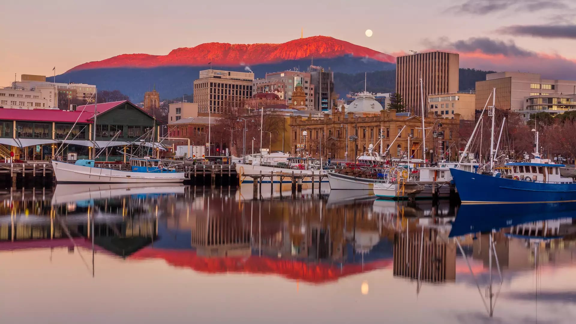 Fishing boats docked in a still harbor reflecting historic stone buildings and a pink mountain under a full moon at dawn in Tasmania.