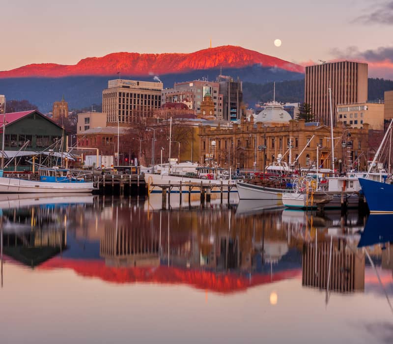 Fishing boats docked in a still harbor reflecting historic stone buildings and a pink mountain under a full moon at dawn in Tasmania.