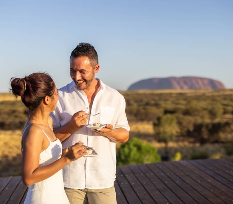 A man in a white shirt and a woman in a white dress stand on a wooden platform, laughing and eating small appetizers, with the distant silhouette of Uluru visible behind them.