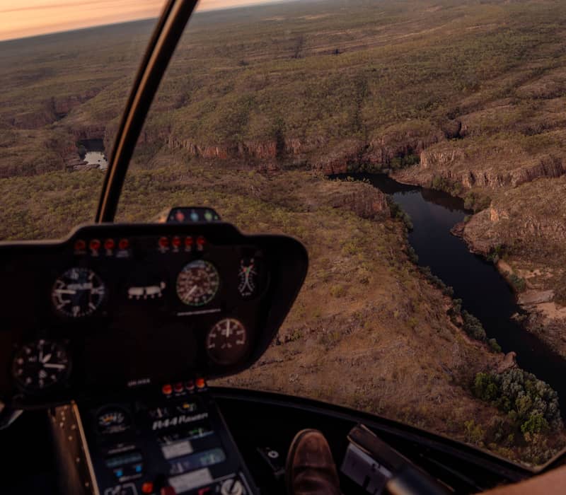 View from inside a helicopter cockpit looking out over the winding Katherine River and rugged gorge landscape at sunset.