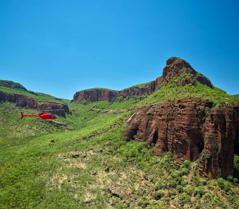 A bright red helicopter flies through a valley beside a massive, green-dusted rocky escarpment under a clear blue sky.