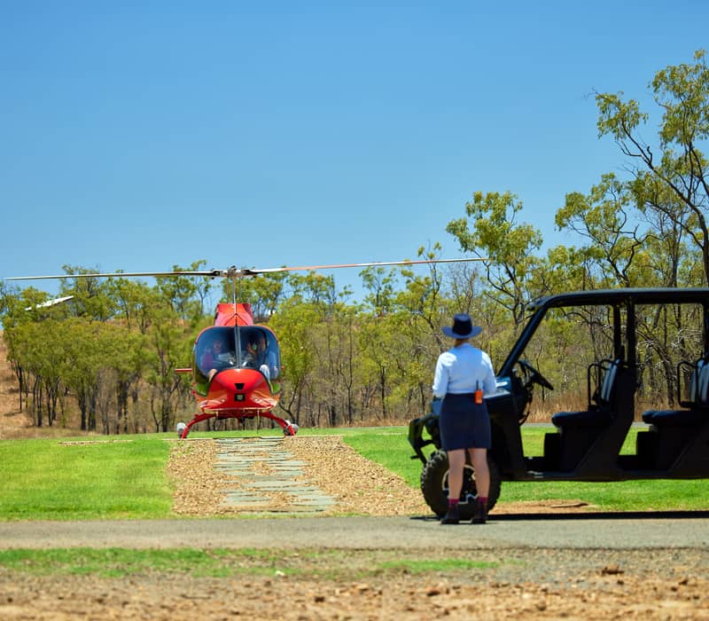 A staff member in a blue shirt and hat stands near an off-road buggy as a red helicopter sits on a grass landing pad under a clear sky.
