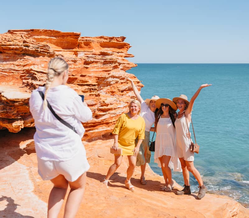Four women posing for a photo on bright red coastal rocks next to a clear blue ocean.