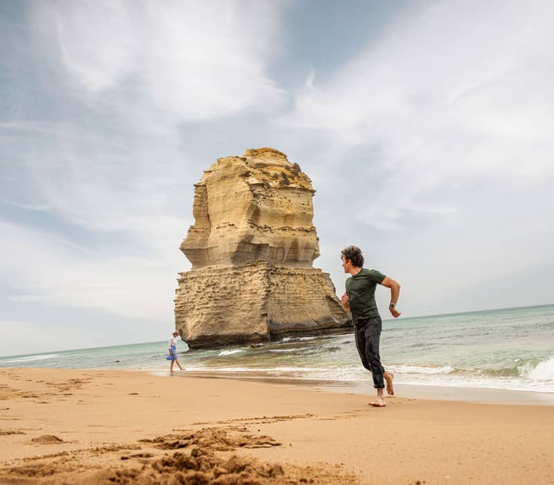 A man running on a sandy beach in front of a giant limestone sea stack under a cloudy sky.