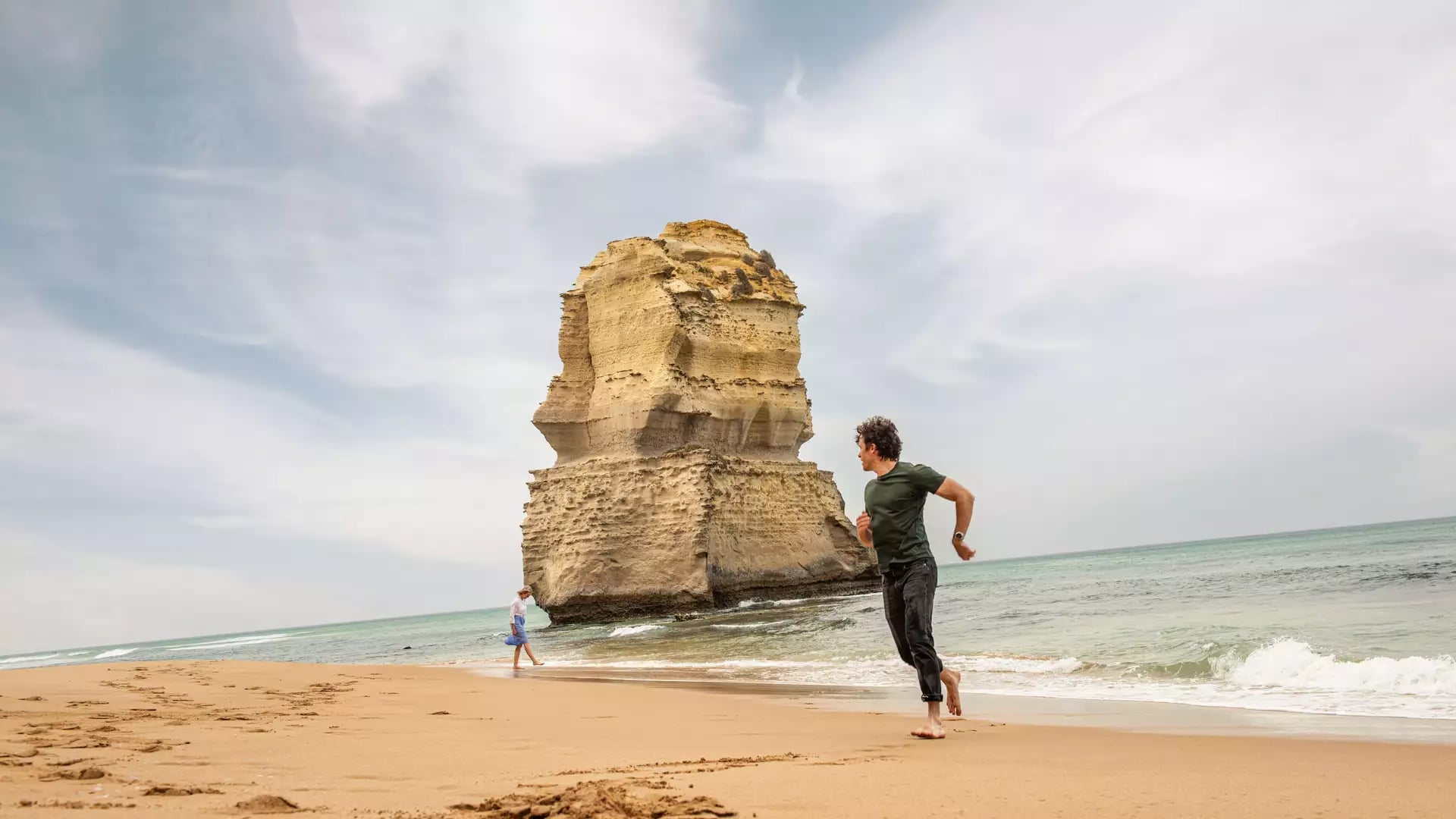 A man running on a sandy beach in front of a giant limestone sea stack under a cloudy sky.