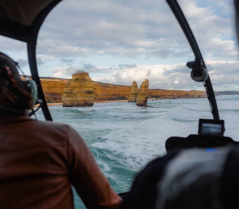 View from inside a helicopter cockpit looking out at the Twelve Apostles rock stacks and crashing waves.
