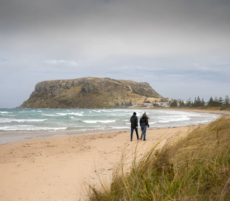 Two people walking on a sandy beach with the large geological formation of The Nut in the distance.