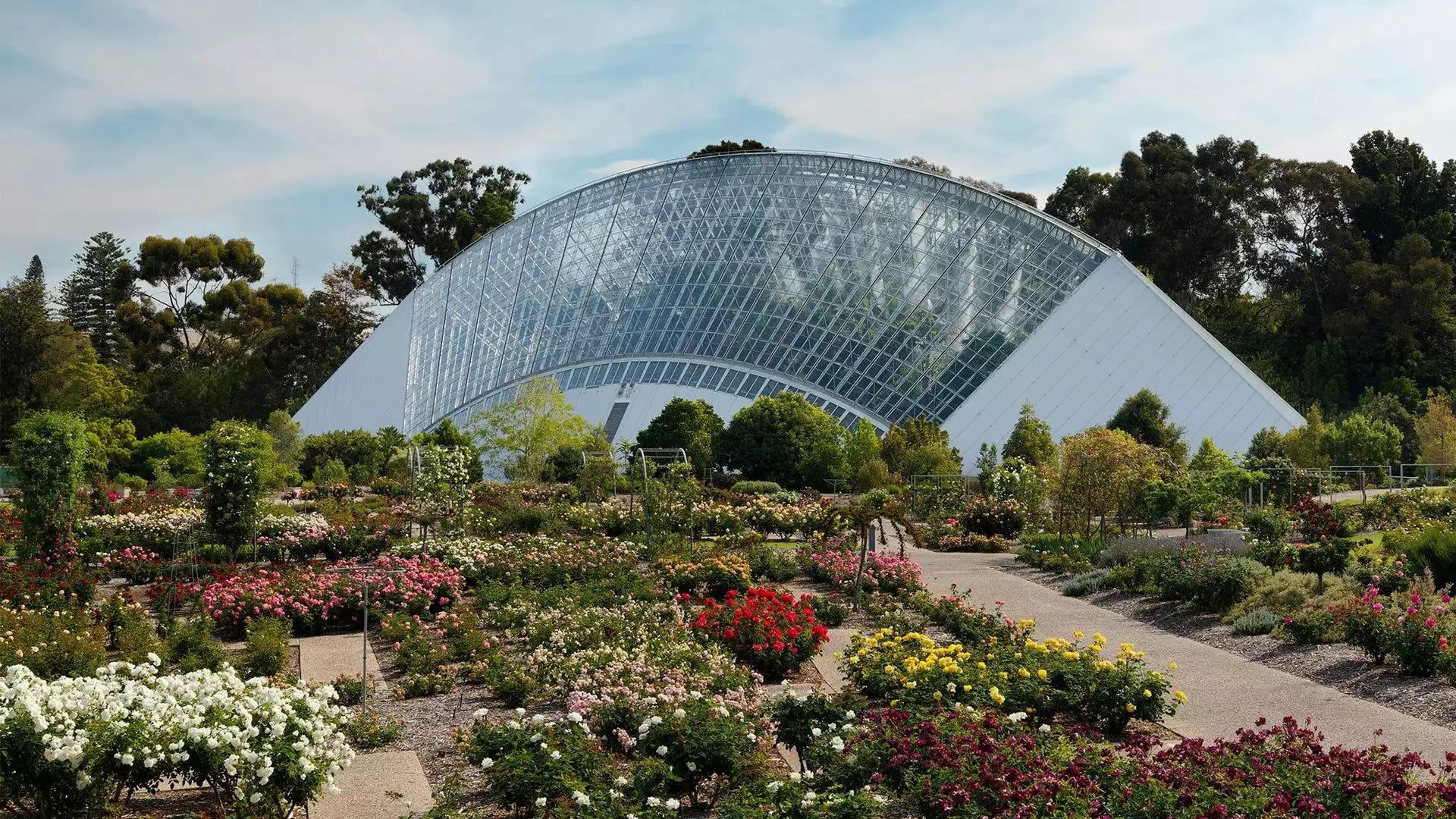 Colorful rose garden with a large glasshouse and trees under a partly cloudy sky