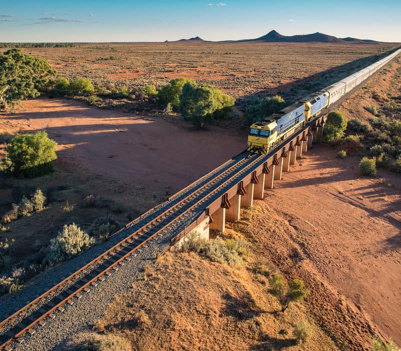 An aerial view of the long blue and yellow Indian Pacific passenger train winding along a curved track through vibrant, rolling emerald-green hills under a bright blue sky.