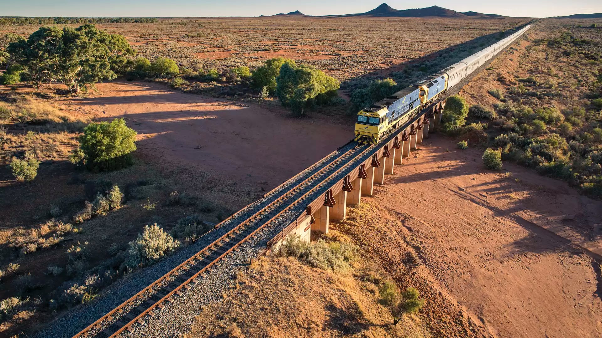 An aerial view of the long blue and yellow Indian Pacific passenger train winding along a curved track through vibrant, rolling emerald-green hills under a bright blue sky.