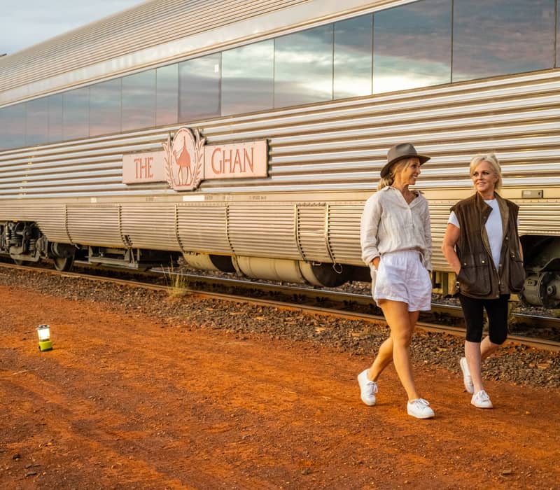 Two women walking on red dirt next to a silver train with "The Ghan" logo during a sunrise stop in Marla.