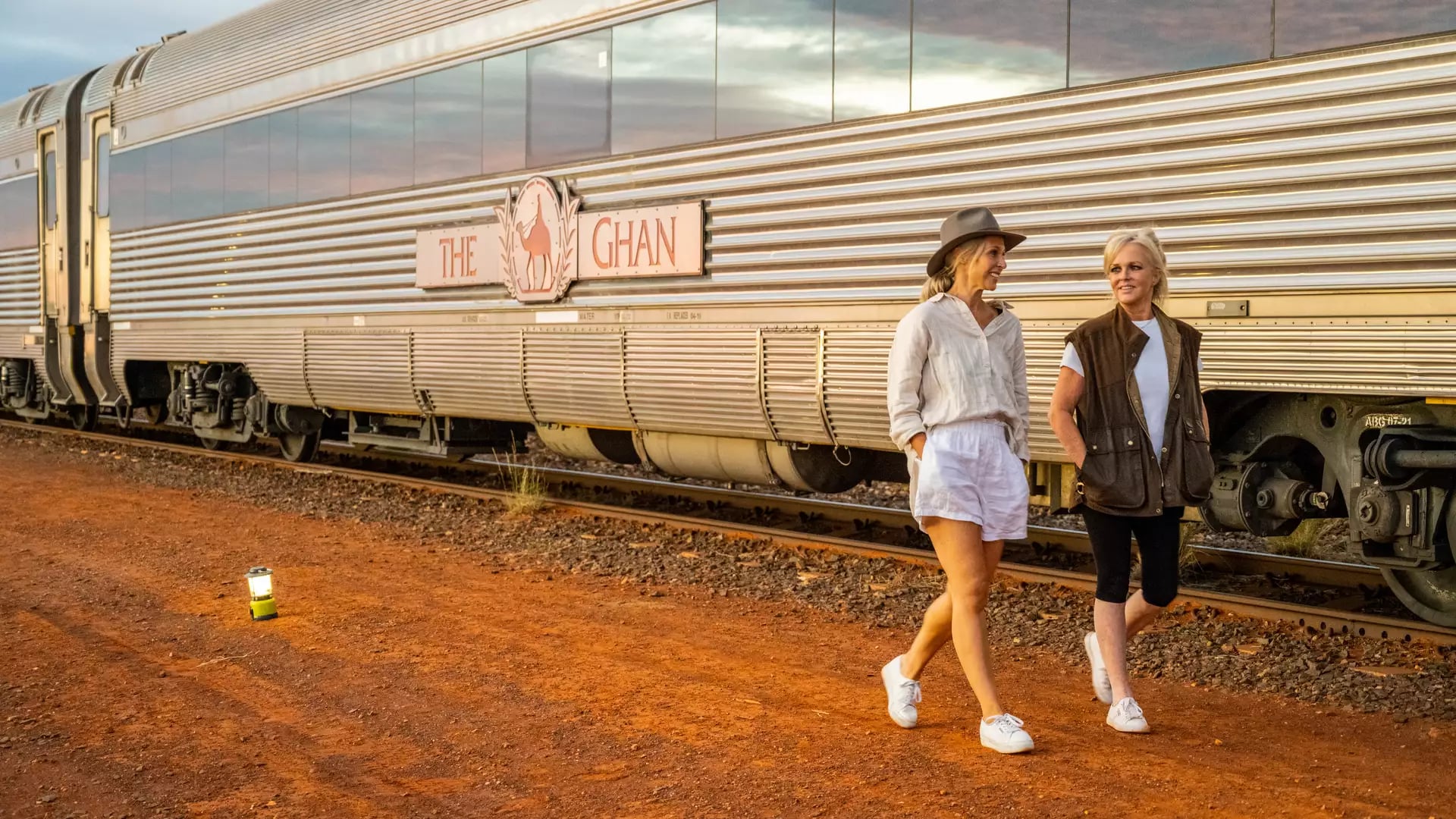 Two women walking on red dirt next to a silver train with "The Ghan" logo during a sunrise stop in Marla.