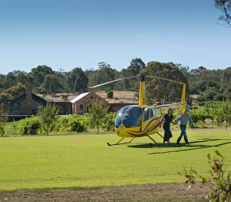 A man and woman walking hand-in-hand away from a yellow helicopter landed on a green lawn in front of heritage stone winery buildings.