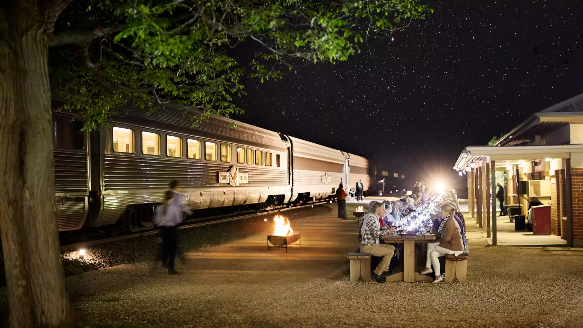 A silver Indian Pacific passenger train is parked alongside a remote station building at Rawlinna, where a large group of guests is seated at long outdoor wooden tables for a communal meal under a cloudy twilight sky.