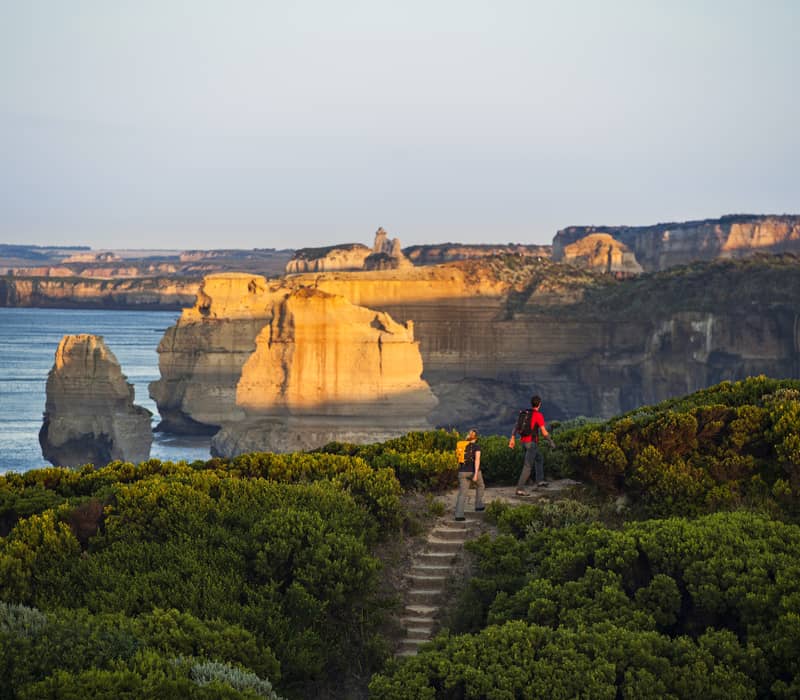 Couple with hiking packs walking up outdoor steps on a green hillside overlooking golden-lit limestone sea stacks.