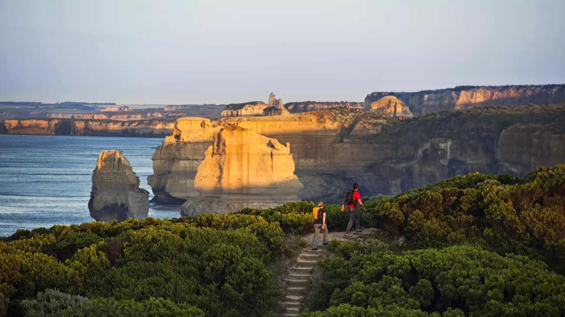 Couple with hiking packs walking up outdoor steps on a green hillside overlooking golden-lit limestone sea stacks.