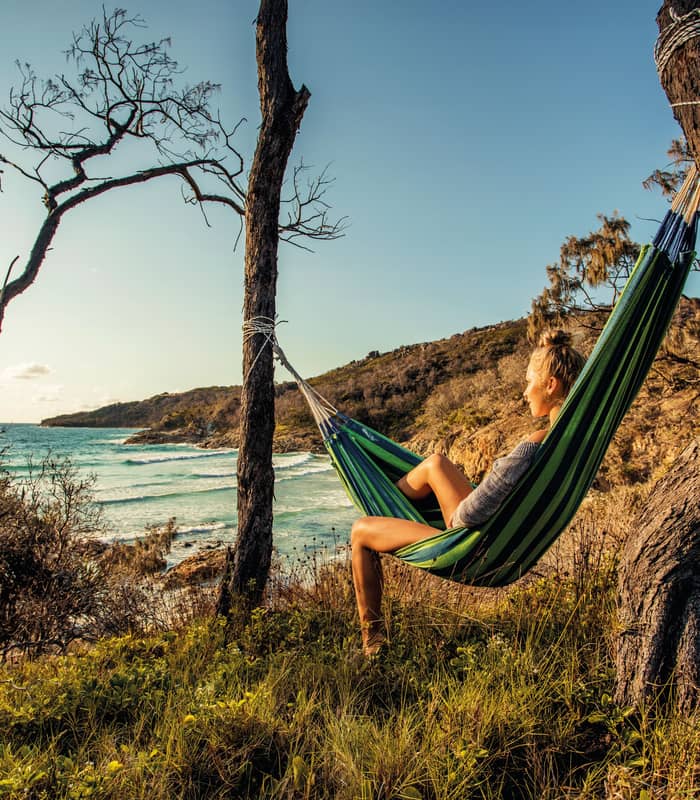 A woman rests in a hammock strung between two trees on a grassy hill, looking out over the turquoise waves and white foam of a Moreton Island beach in Queensland.