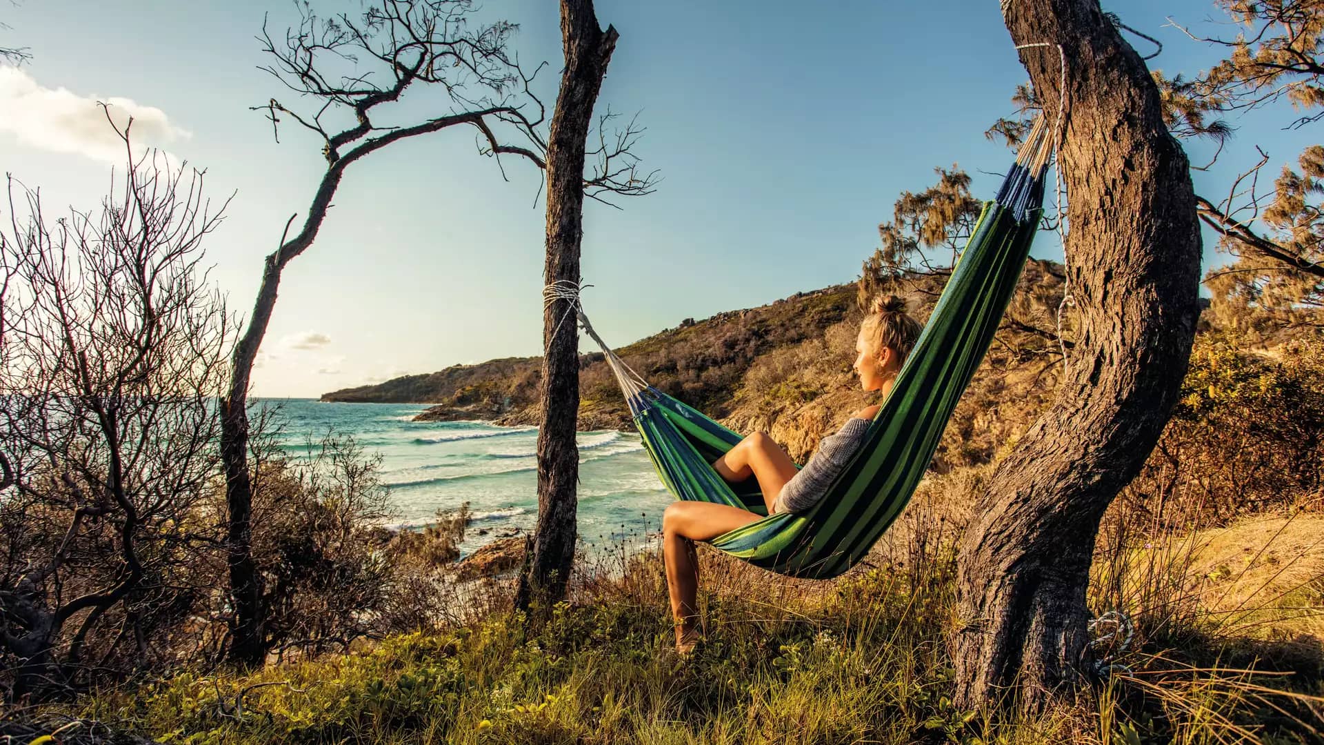 A woman rests in a hammock strung between two trees on a grassy hill, looking out over the turquoise waves and white foam of a Moreton Island beach in Queensland.