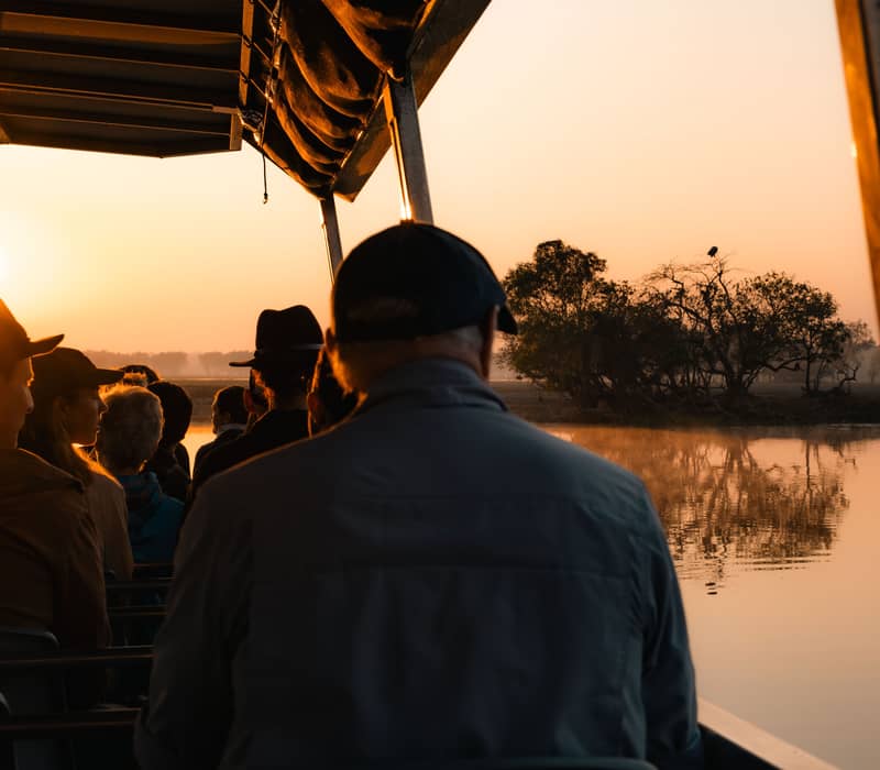 Silhouetted passengers on a tour boat watch a bright golden sunrise over the calm, misty waters of a billabong in Kakadu National Park.