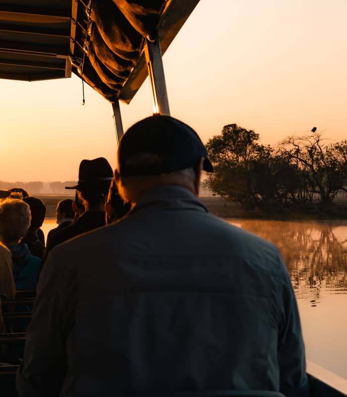 Silhouetted passengers on a tour boat watch a bright golden sunrise over the calm, misty waters of a billabong in Kakadu National Park.