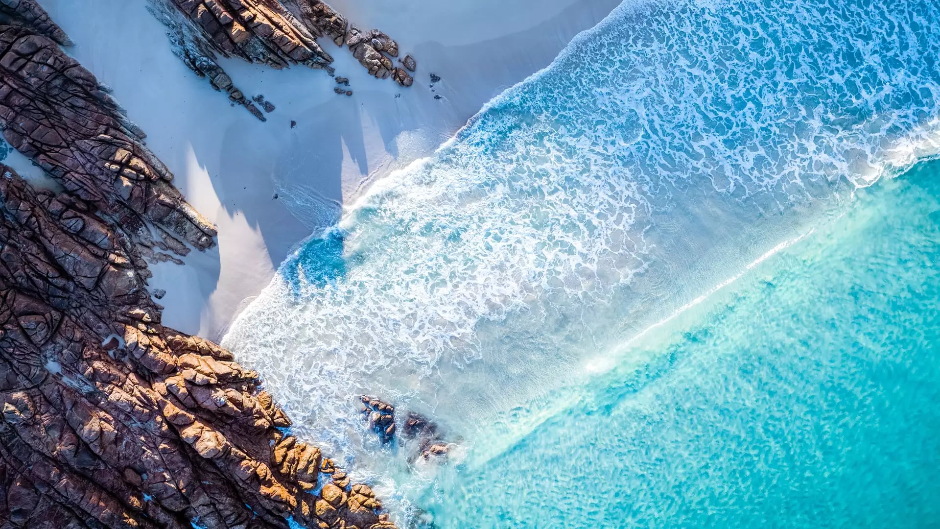 A dramatic top-down aerial view captures the white foam of the Indian Ocean crashing against the ancient red-tinted granite of Wyadup Rocks in Yallingup.