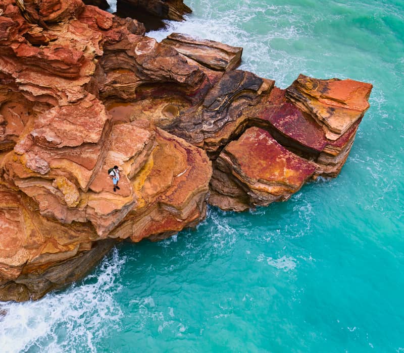 Aerial perspective of a person standing on jagged red rock cliffs overlooking vibrant bright blue ocean water.