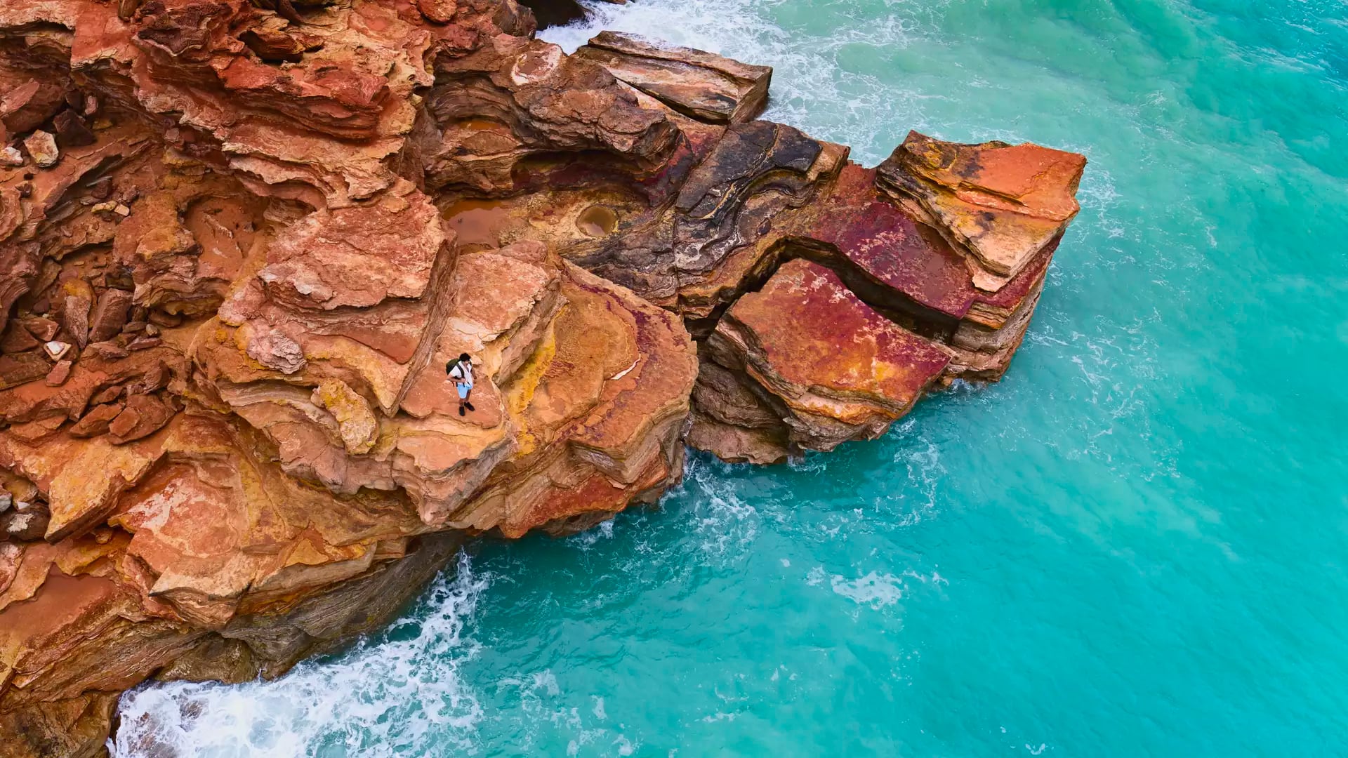 Aerial perspective of a person standing on jagged red rock cliffs overlooking vibrant bright blue ocean water.