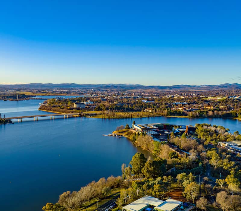 High-angle landscape of Lake Burley Griffin and the Canberra city skyline under a clear blue sky.