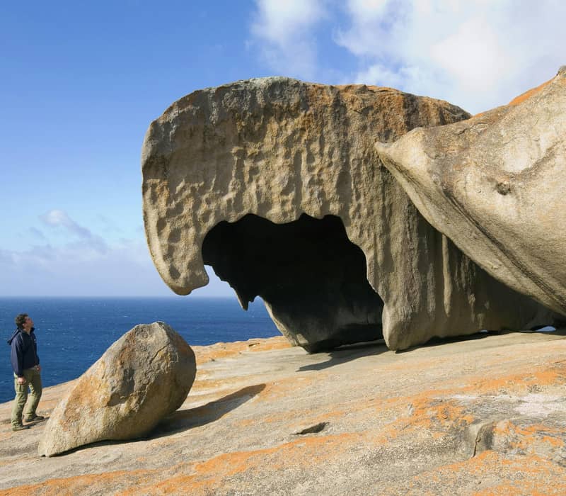 A man standing next to massive, uniquely shaped granite boulders overlooking the blue ocean at Remarkable Rocks, Kangaroo Island.