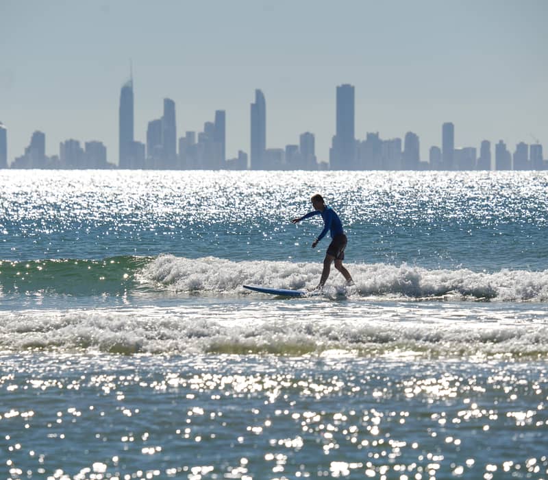 A person surfing on a sparkling blue ocean with a silhouetted city skyline of high-rise buildings in the background.