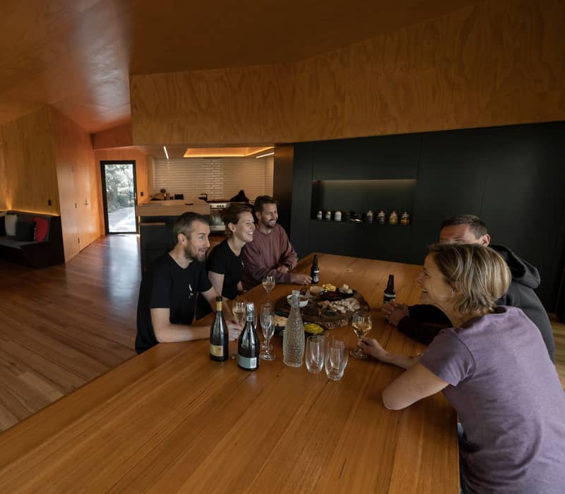People sitting around a large timber table in a modern plywood-lined cabin with a roaring fireplace.