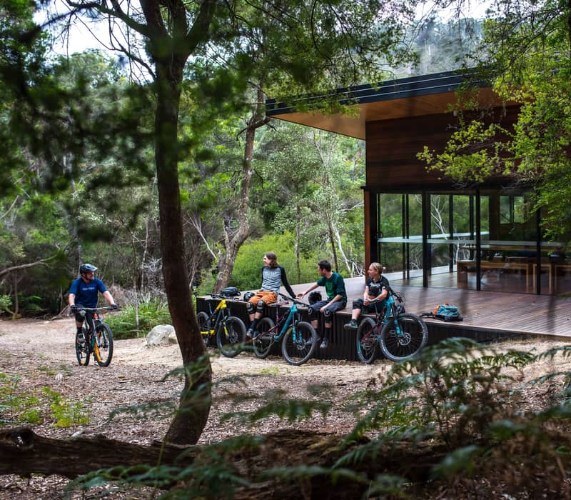 Cyclists resting on a wooden deck outside a contemporary black and timber building in the woods.